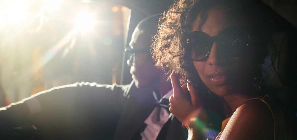 Woman in round sunglasses with a labret stud below her lip, lit by camera flash beside a man in a bow tie