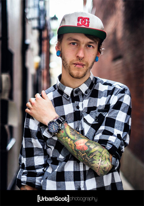 Man in a snapback cap with blue plugs in stretched ears, a septum ring, snake bite rings and a tattooed forearm