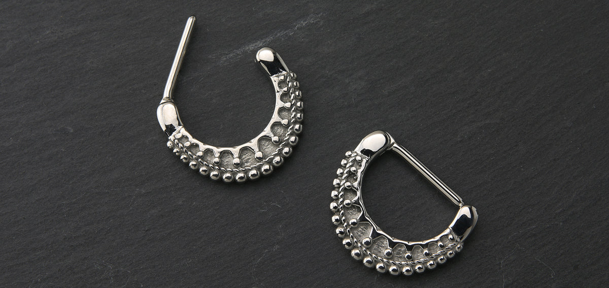 Two beaded silver-tone nipple clickers, one open and one closed, on dark slate
