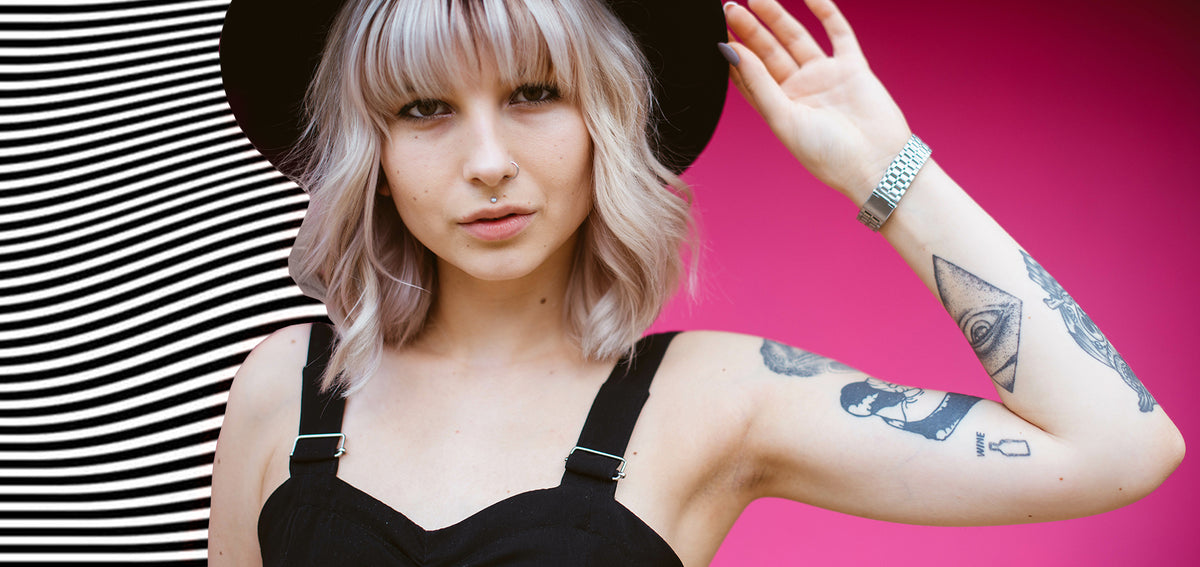 Woman with a platinum bob and black hat wearing a thin nostril hoop and a medusa stud, her tattooed arm raised
